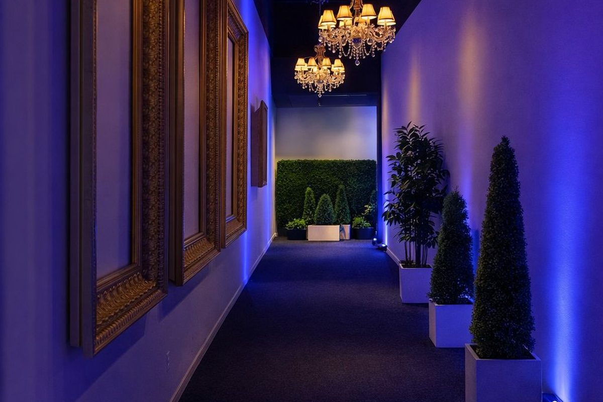 cases an elegantly lit modern corridor featuring ornate gold-framed artwork on one wall, hanging crystal chandeliers with…