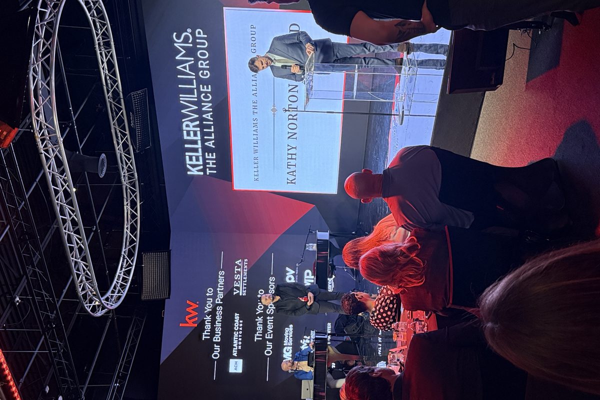 This image captures a professional corporate event for Keller Williams The Alliance Group, featuring a man speaking at a…