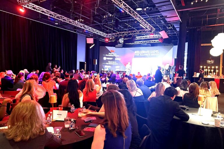 This image captures a professional gala or awards event featuring multiple attendees seated at formal dining tables facing a…