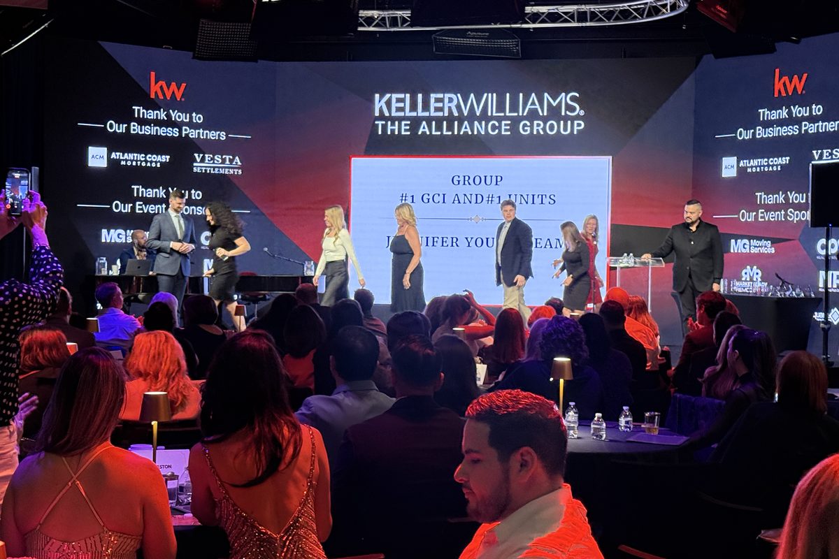 This image captures a professional Keller Williams real estate conference or gala event, featuring a panel of speakers on…