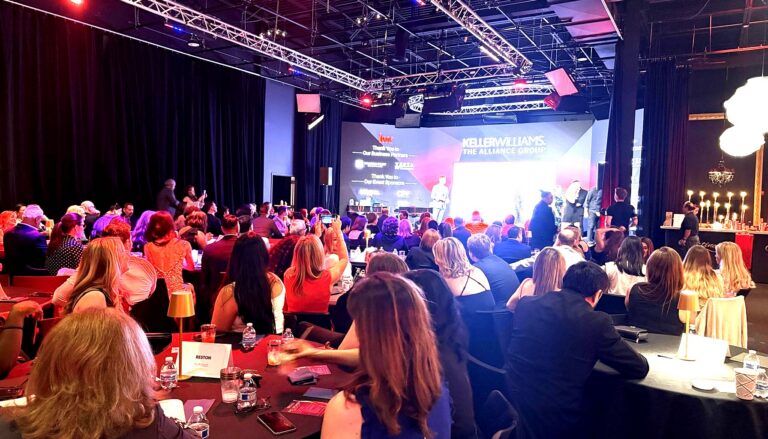 This image captures a professional gala or awards event featuring multiple attendees seated at formal dining tables facing a…
