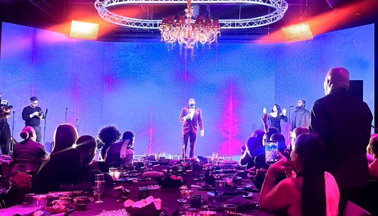 A glamorous indoor gala or formal event features live entertainment on stage with a performer in a burgundy suit…