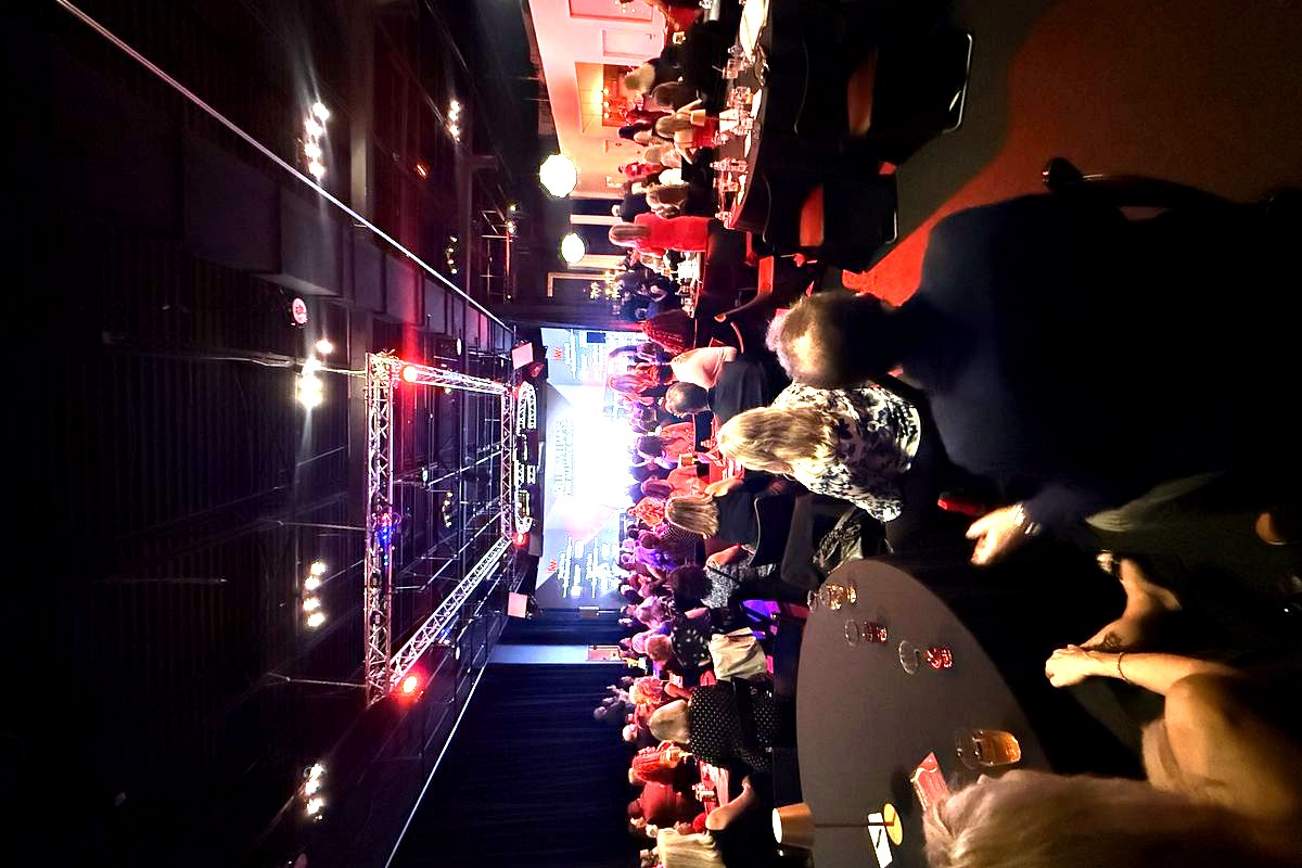 This image captures a large indoor gala or corporate event with hundreds of attendees seated at round tables in a darkly lit…