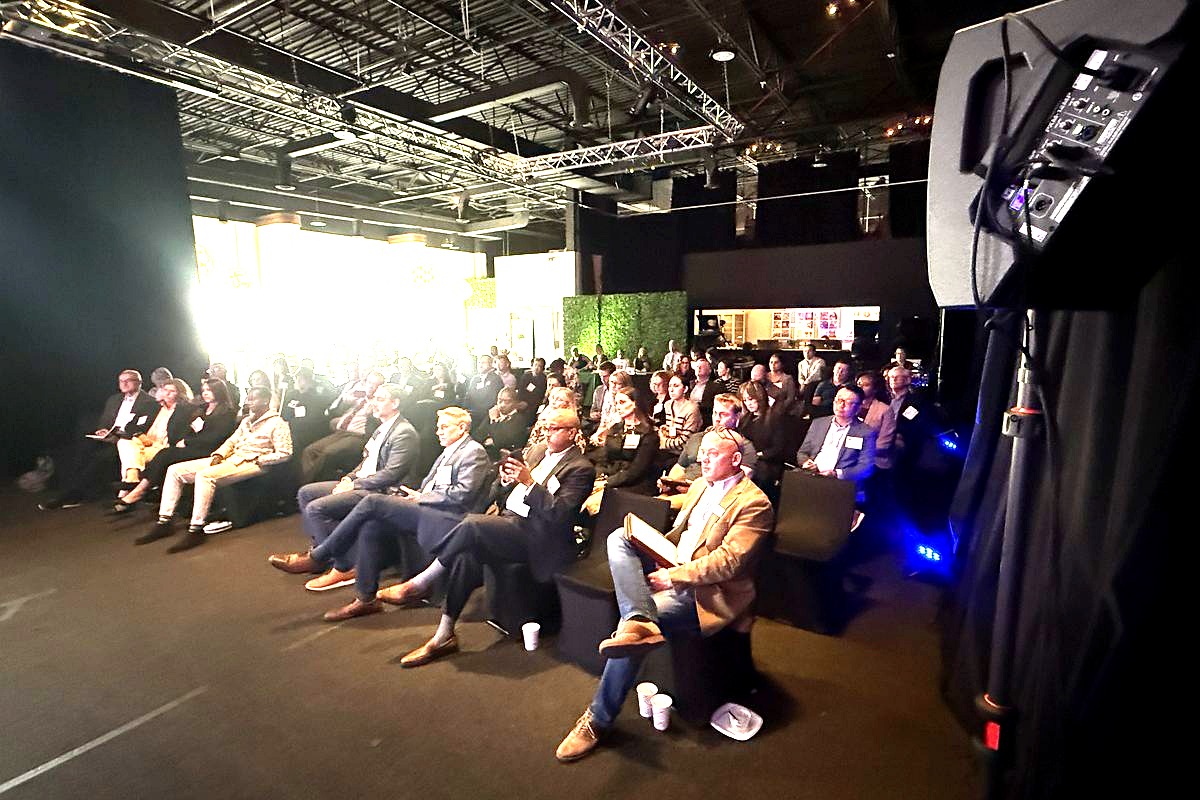This image captures a professional conference or corporate event in a modern warehouse-style venue, with a large audience of…