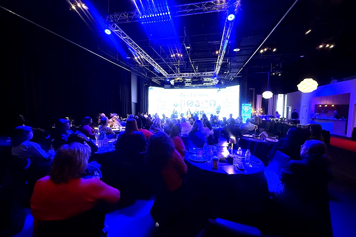 This image captures a professional conference or gala event with hundreds of attendees seated at round tables in a modern…