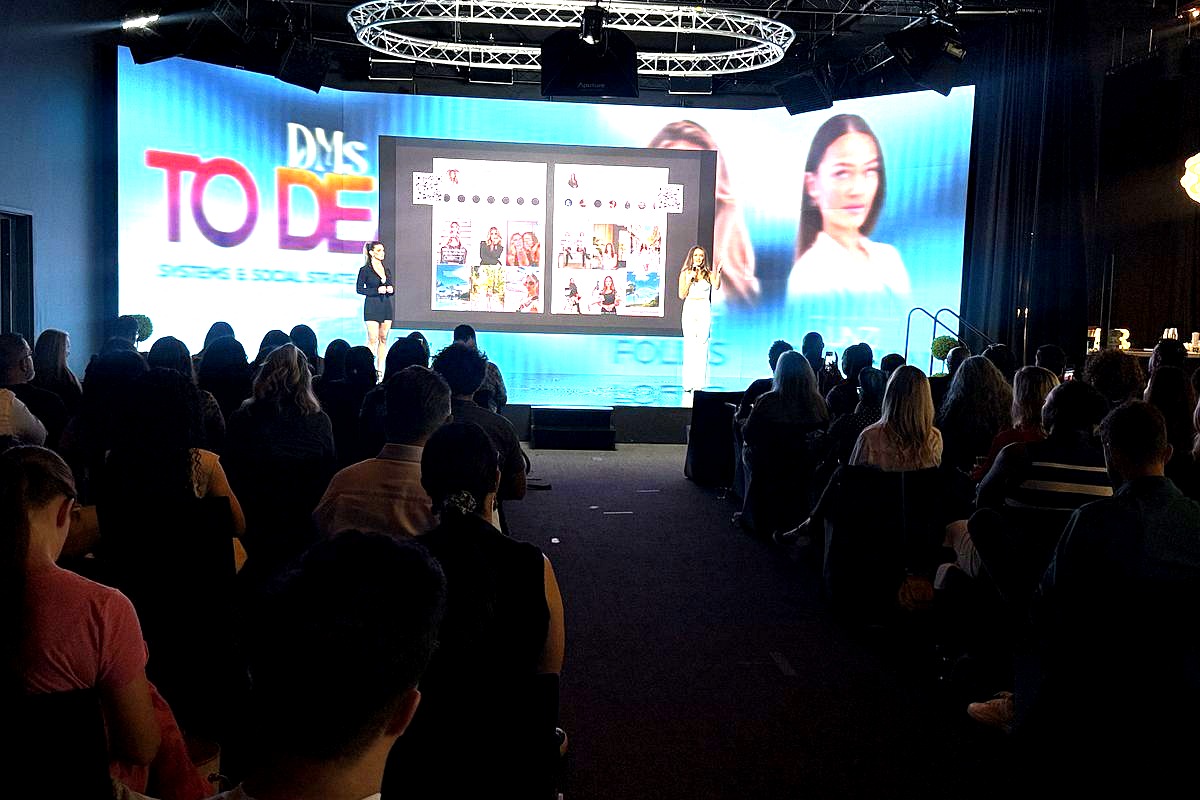A large audience watches a professional presentation on stage where two women present social media content displayed on a…