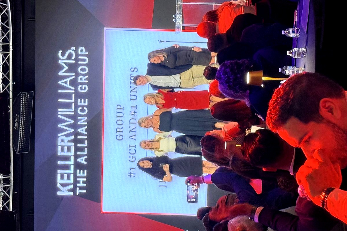 A professional awards or recognition event shows six team members from Keller Williams The Alliance Group standing on stage…
