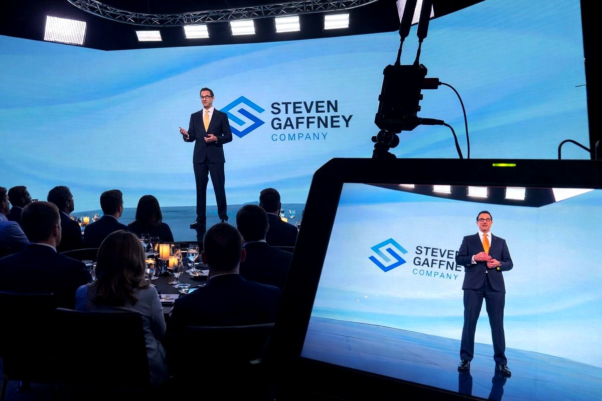 A professional business presentation featuring a man in a dark suit and yellow tie speaking on stage at what appears to be a…