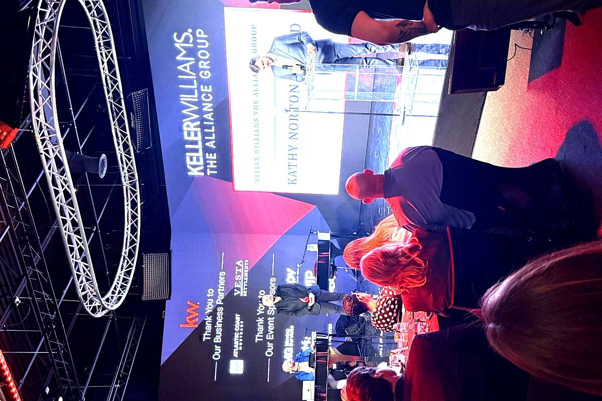 This image captures a professional corporate event for Keller Williams The Alliance Group, featuring a man speaking at a…