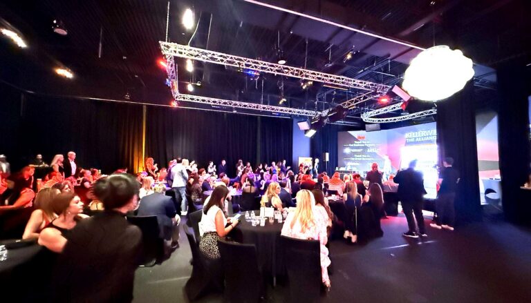 This image captures an elegant indoor gala or formal event with hundreds of guests seated at black tables in a…