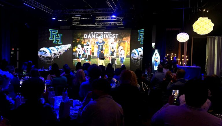 This image captures an indoor awards ceremony honoring Dane Rivest with the JV Coaches Award, featuring a large projection…