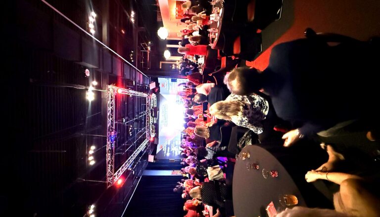This image captures a large indoor gala or corporate event with hundreds of attendees seated at round tables in a darkly lit…