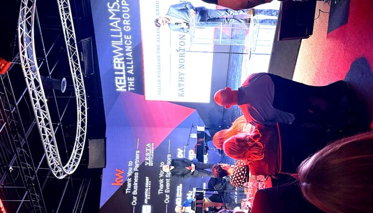 This image captures a professional corporate event for Keller Williams The Alliance Group, featuring a man speaking at a…